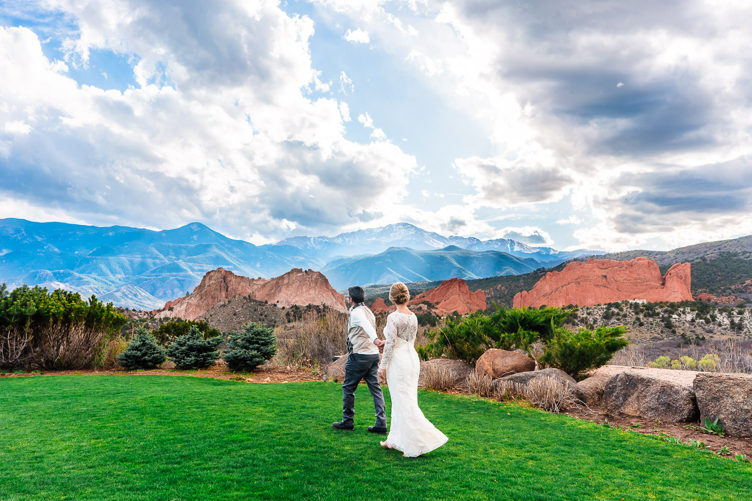 Newlyweds in wedding attire hold hands and walk along a grassy area in Garden of the Gods resort with distant views of the Garden of the Gods park and the Rocky Mountains.