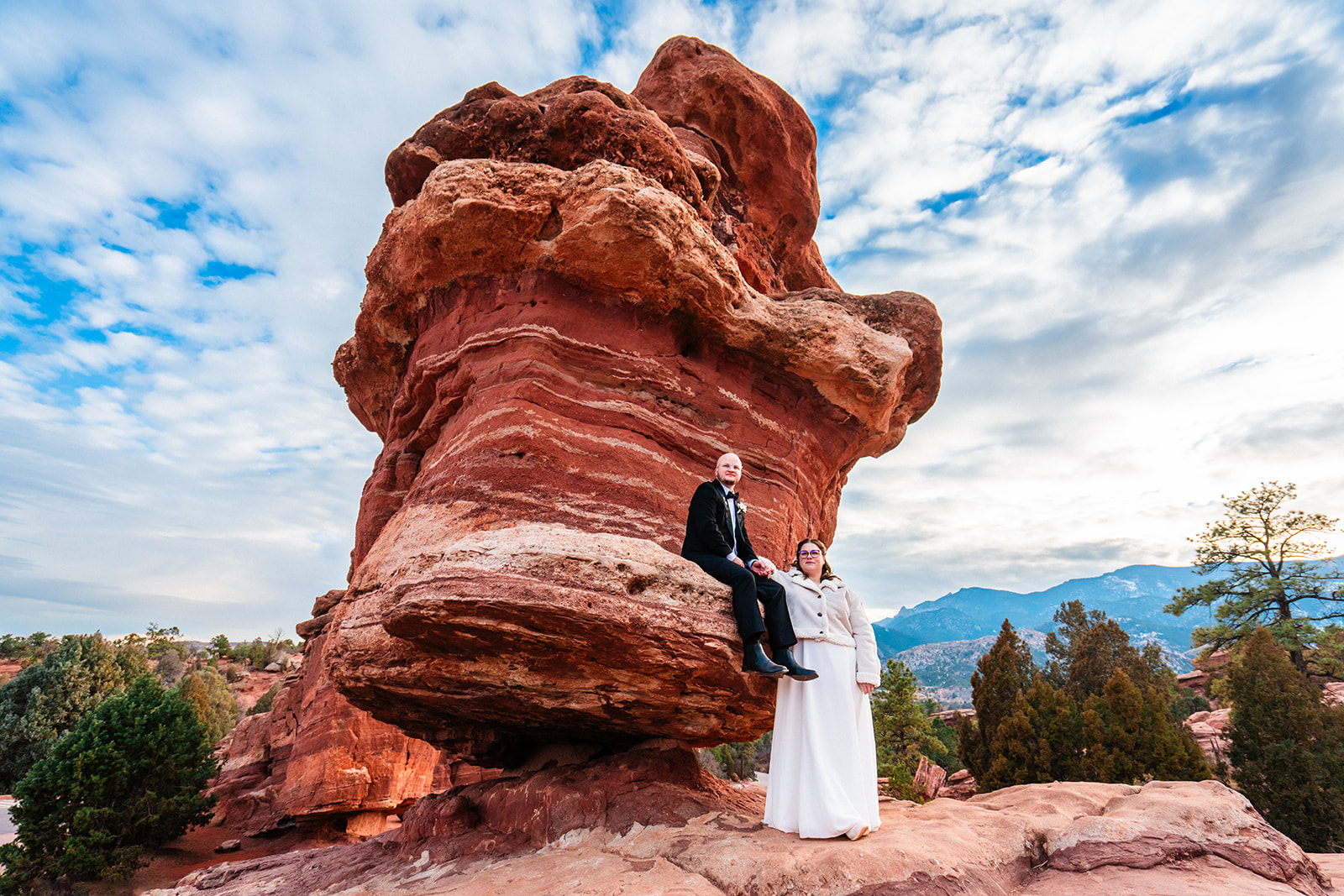 Dapper newlyweds hang out on the breathtaking Balanced Rock at Garden of the Gods during their elopement under a blue sky with clouds.
