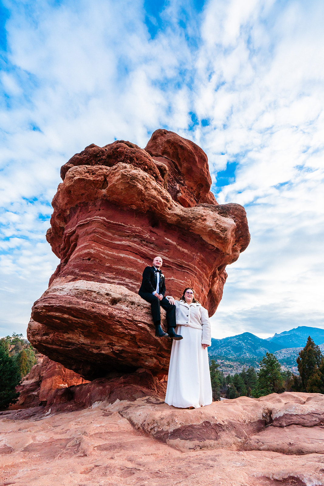 A bride and groom sit upon Balanced Rock during their Garden of the Gods elopement under a blue sky with white clouds.