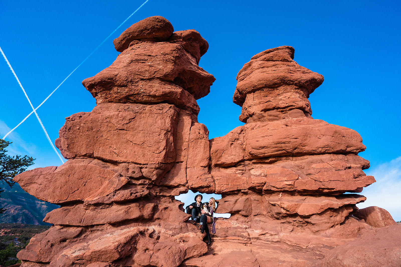 A cute couple sits with their dog between towering red rock formations at Siamese Twins under a blue sky at Garden of the Gods.