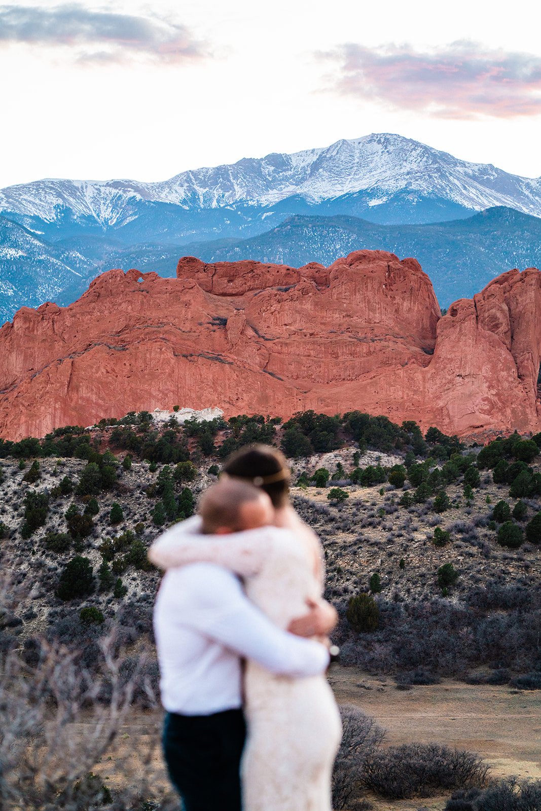 Newlyweds embrace in front of a red rock formation called the kissing camels at the Mesa overlook during their Garden of the Gods elopement with snowy mountains in the background.