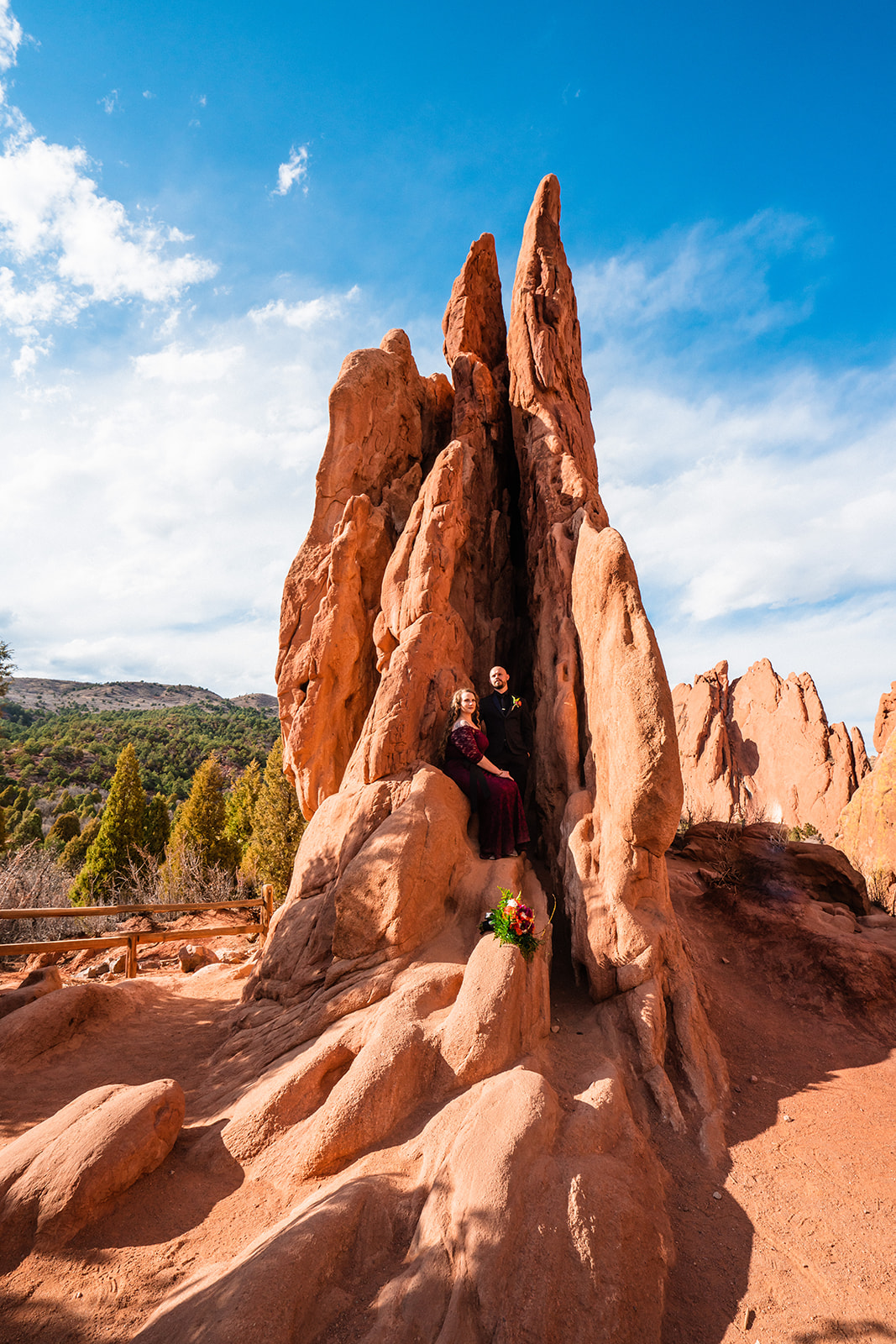 A bride in a burgundy elopement dress explores Three Graces Plaza with her groom beneath a bright blue sky during their Garden of the Gods elopement.