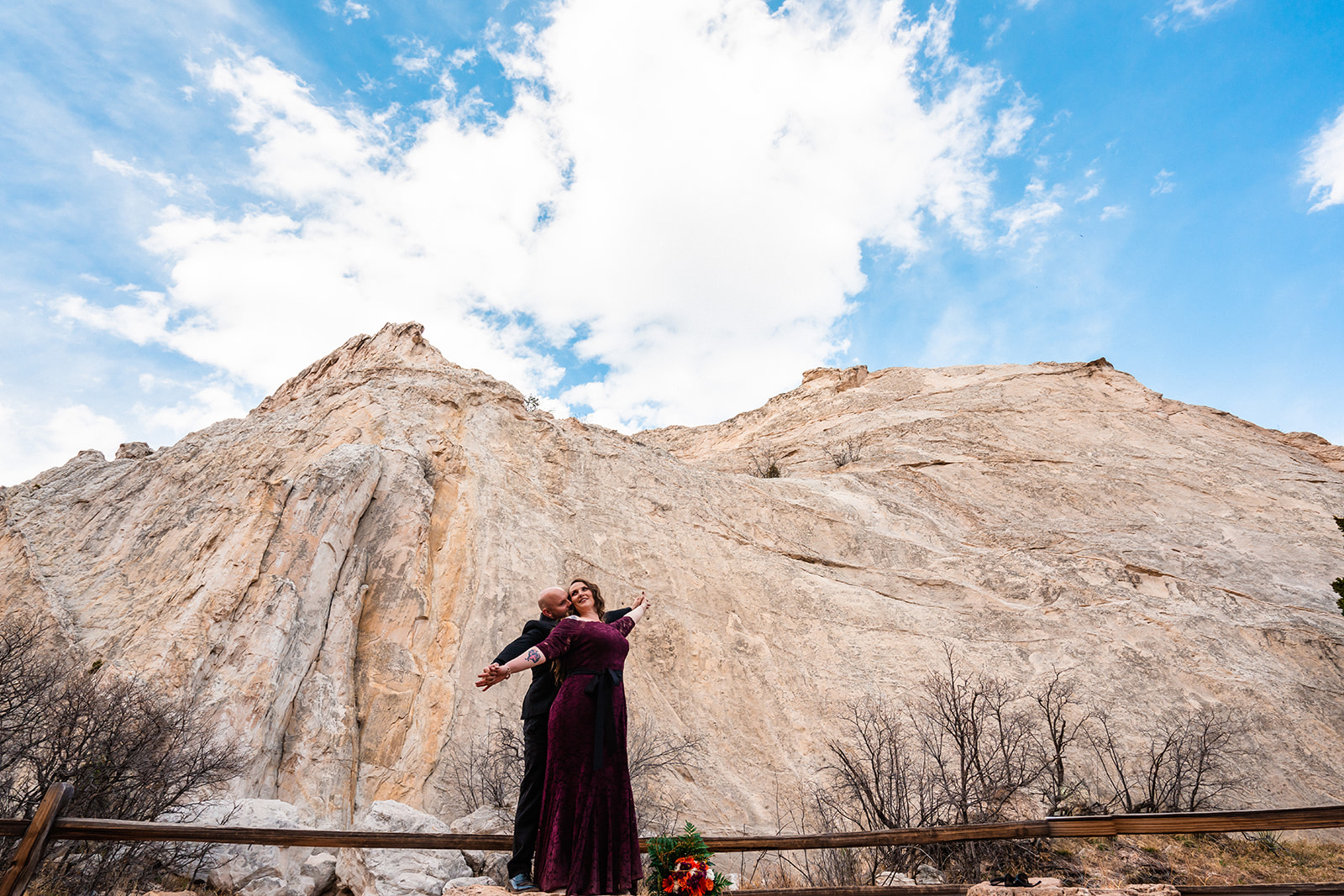 Newlyweds in formal attire embrace at the base of White Rock beneath blue and cloudy skies during their Garden of the Gods elopement in Colorado Springs.