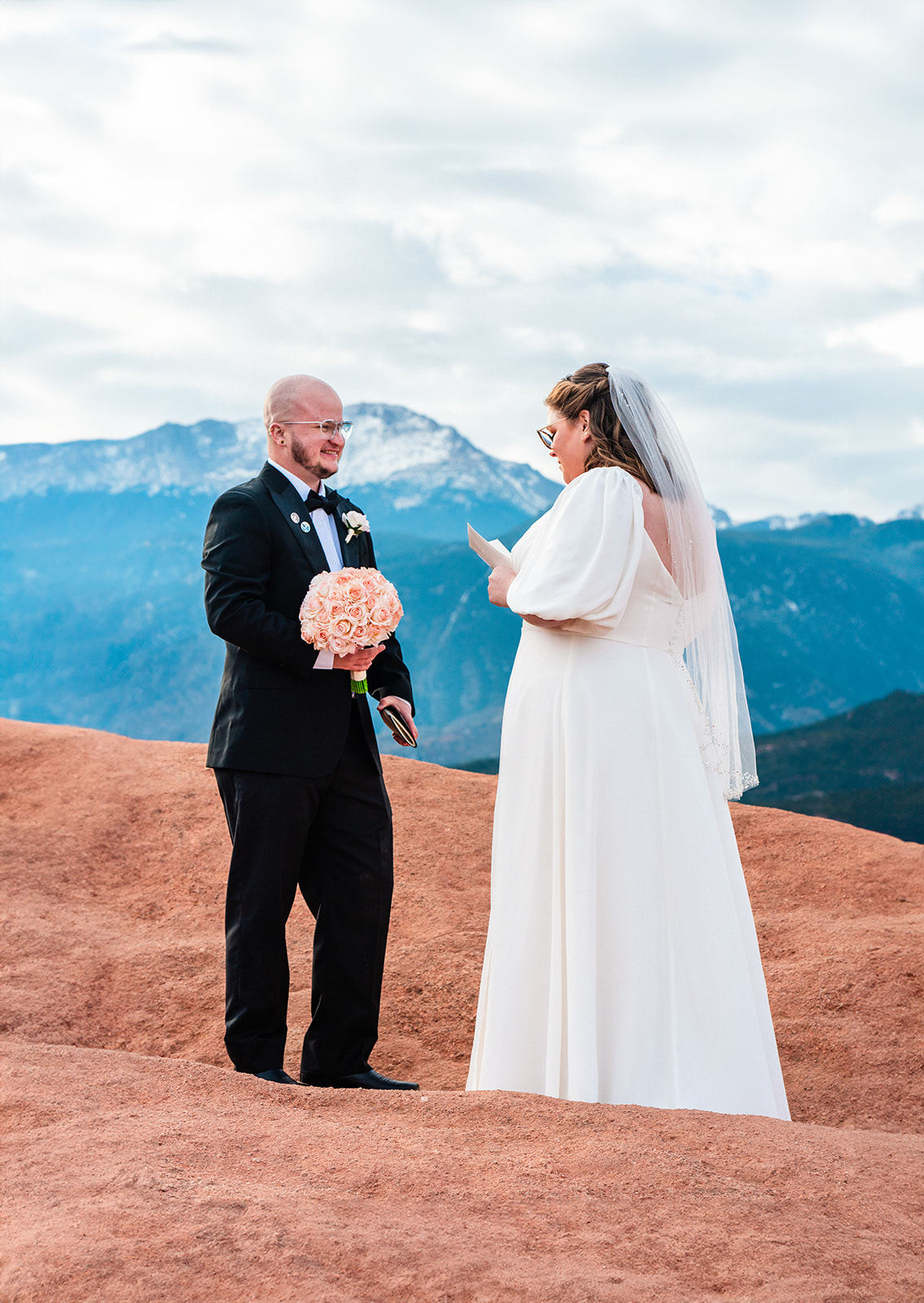 A bride and groom exchanging vows outdoors at High Point in  Garden of the Gods, with a mountainous backdrop, standing on red earth. The groom is bald, and both are smiling, holding hands.